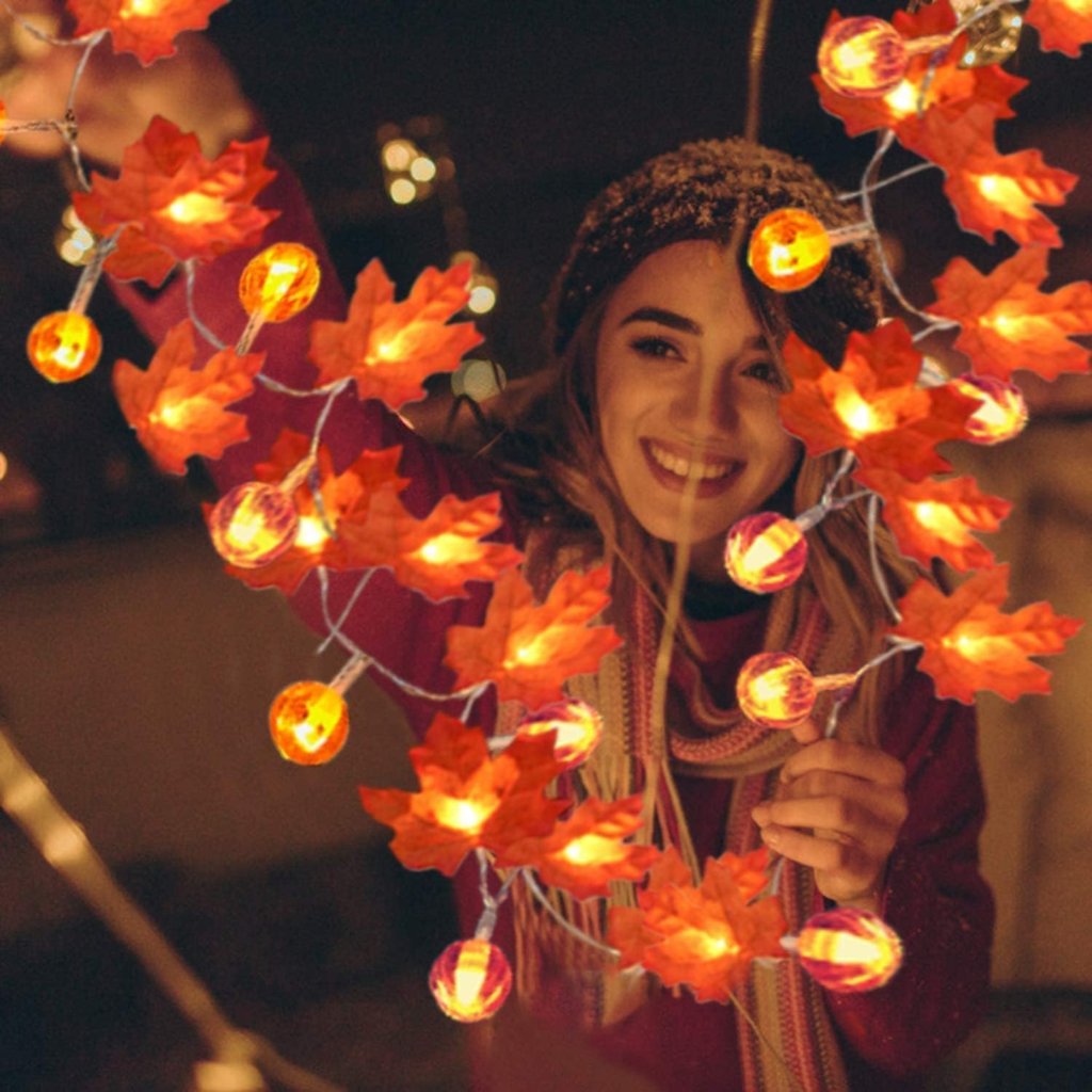 En la imagen, se puede sentir la calidez de una joven que sonríe alegremente. Ella está disfrutando el momento mientras coloca una guirnalda de luces, adornada con hojas de otoño, que evoca una atmósfera acogedora y festiva. Su alegría y la calidez de la escena transmiten una sensación de felicidad y conexión con la belleza de la temporada.